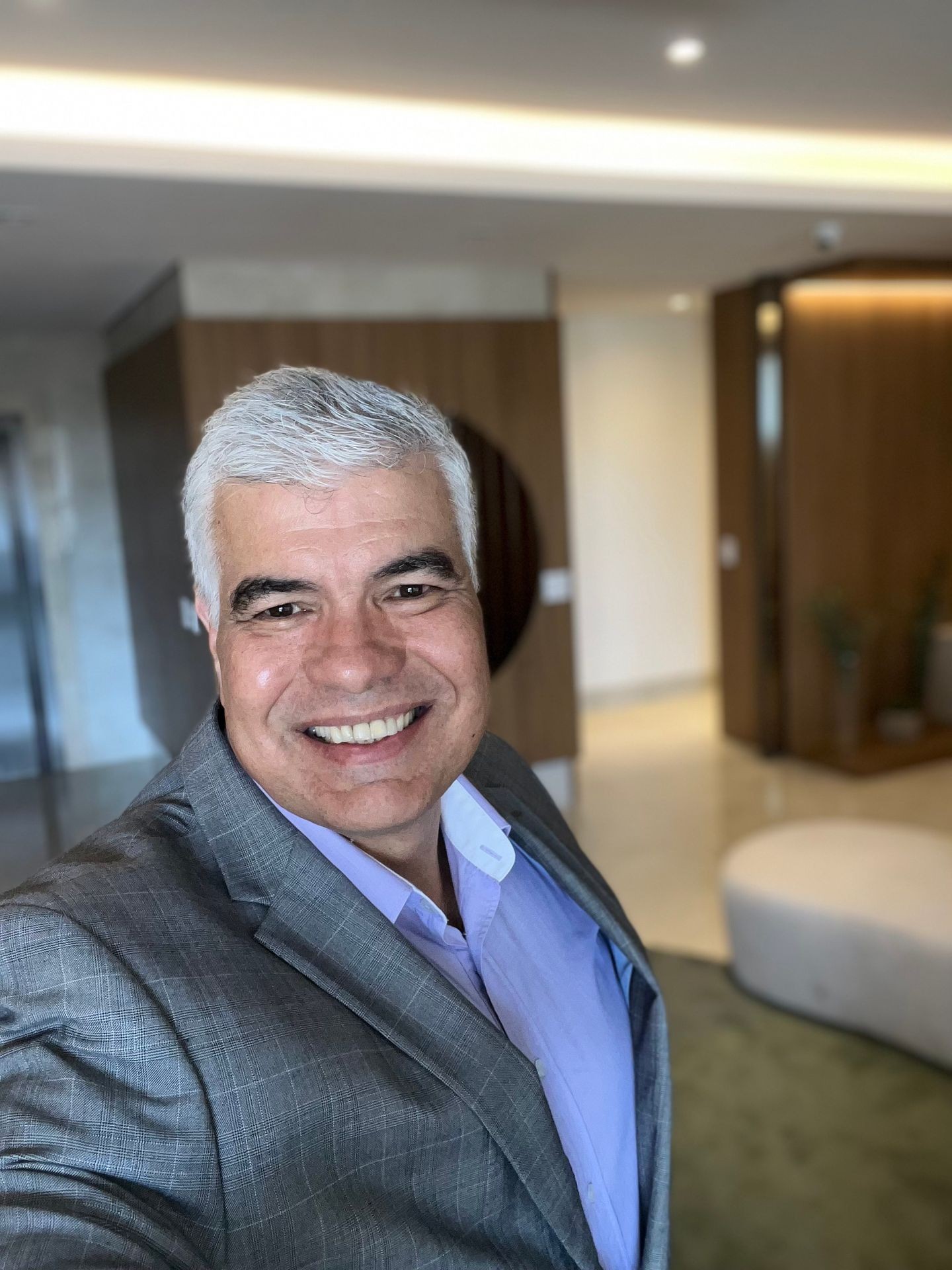 Man in a grey suit and light purple shirt smiling in a modern, well-lit office lobby.