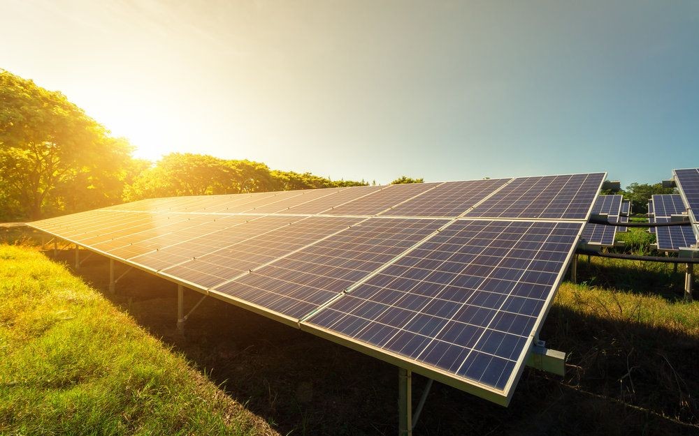 Solar panels installed in a field, absorbing sunlight with trees and a bright sun in the background.